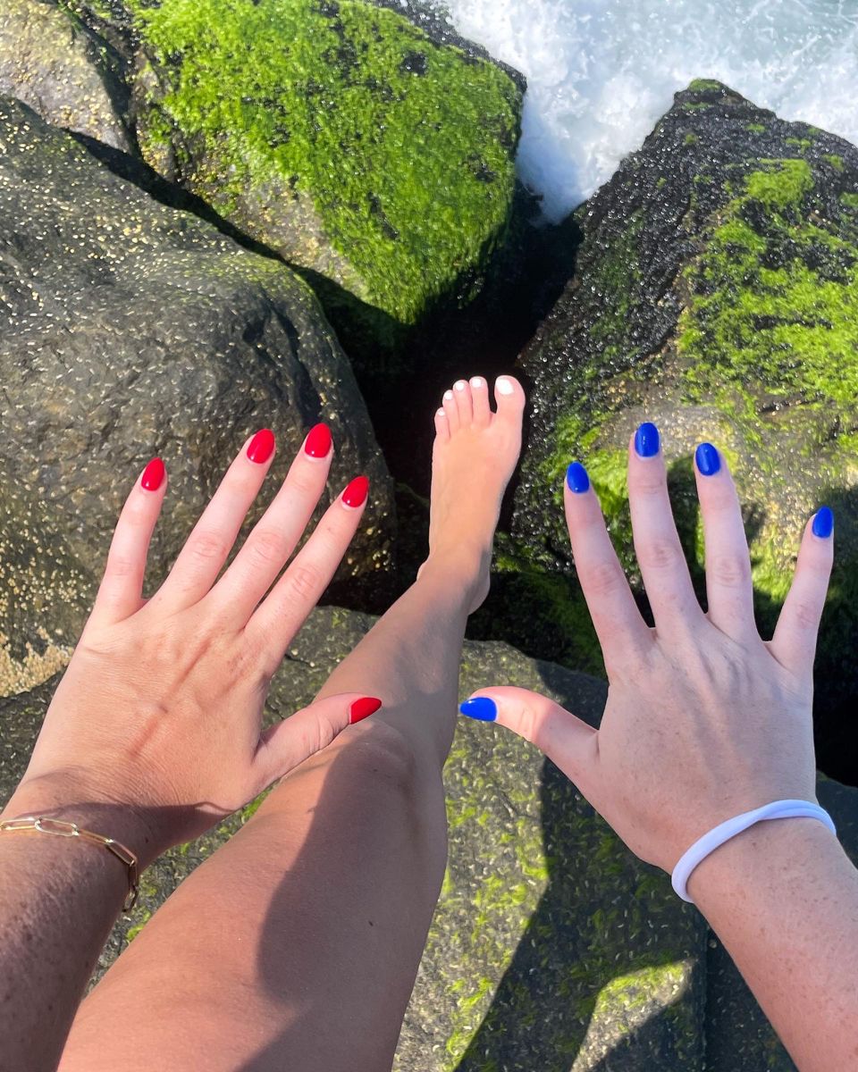Two hands photographed outdoors on moss-covered rocks, one with solid red almond nails and the other with solid cobalt blue almond nails, with bare feet showing white toenails in the background.