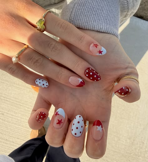 Short almond nails featuring a mix of dark red solid nails, white nails with dark red polka dots, nude nails with a dark red star French tip, and a red and white polka dot accent nail, photographed with gold rings.