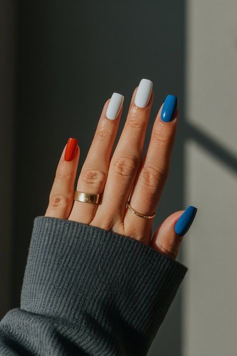 Long square nails with each nail painted a single block colour in the patriotic palette, featuring orange-red, white, and cobalt blue across the fingers, photographed against a dark background.