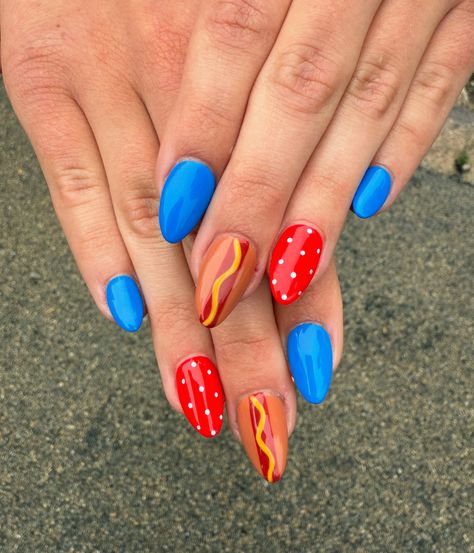 Medium almond nails featuring solid cobalt blue on most fingers with red accent nails, white polka dot details, and a playful hot dog nail art design on two accent fingers.