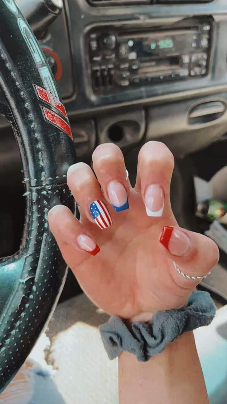 Short coffin nails featuring a mix of patriotic French tip designs including a red tip, white tip, blue V-shaped tip, and a detailed American flag accent nail, photographed in a car.