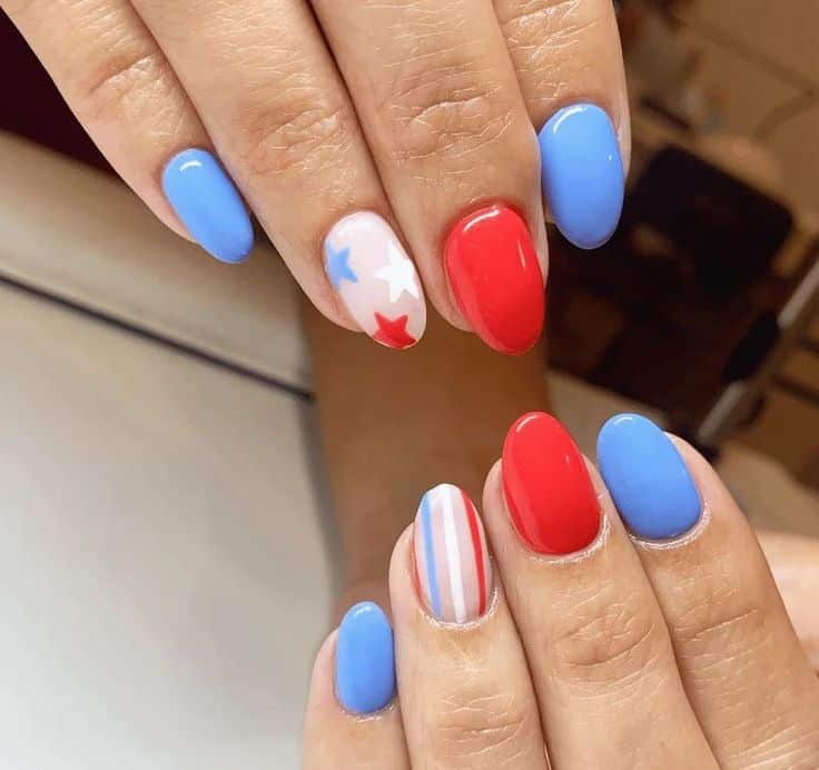 Short oval nails painted in solid blue and red with one white nail featuring red and blue stars and another with red and white stripes, shown on two hands.