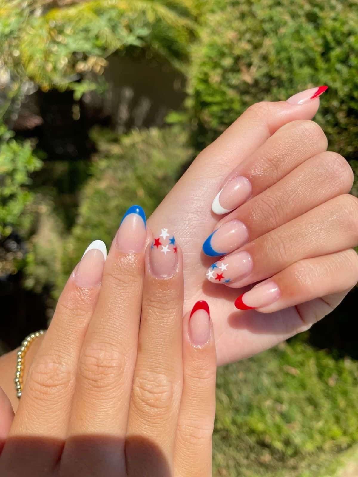 Medium almond nails with a sheer nude base featuring individual French tips in red, white, and blue and scattered red, white, and blue stars across the nail beds, photographed outdoors against greenery.