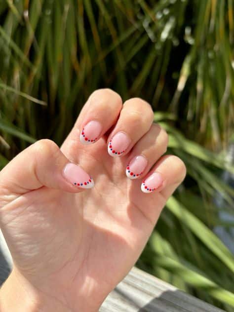 Short oval nails with a natural nude base and white French tips, each smile line decorated with a delicate row of alternating small red and blue dots, photographed outdoors with palm trees in the background.