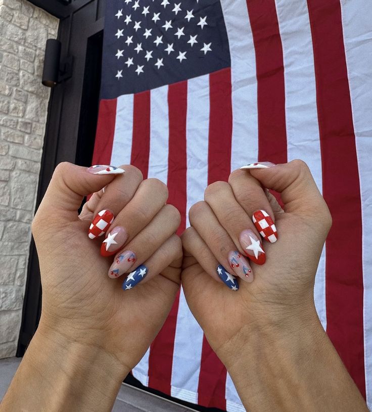 Medium almond nails featuring a patriotic nail set with red and white checkered nails, a blue nail with white stars, a nude nail with red cherry details, and a small red heart accent, photographed in front of an American flag.