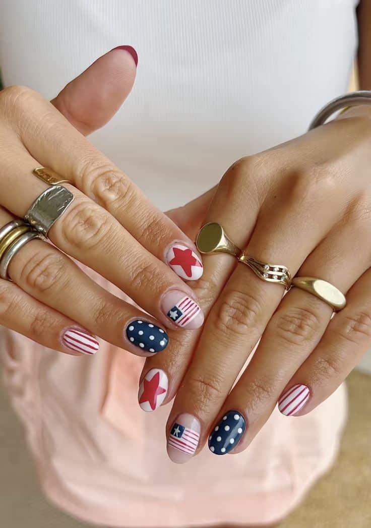 Short oval nails displaying a full patriotic nail art set with individual designs including red stars on a nude base, navy polka dots, red and white stripes with a small flag detail, and red and blue accents on each finger.