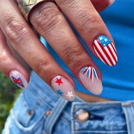 Medium almond nails featuring an American flag design on one nail, a red and white starburst fireworks nail, scattered star nails on a nude base, and a navy star outline nail.