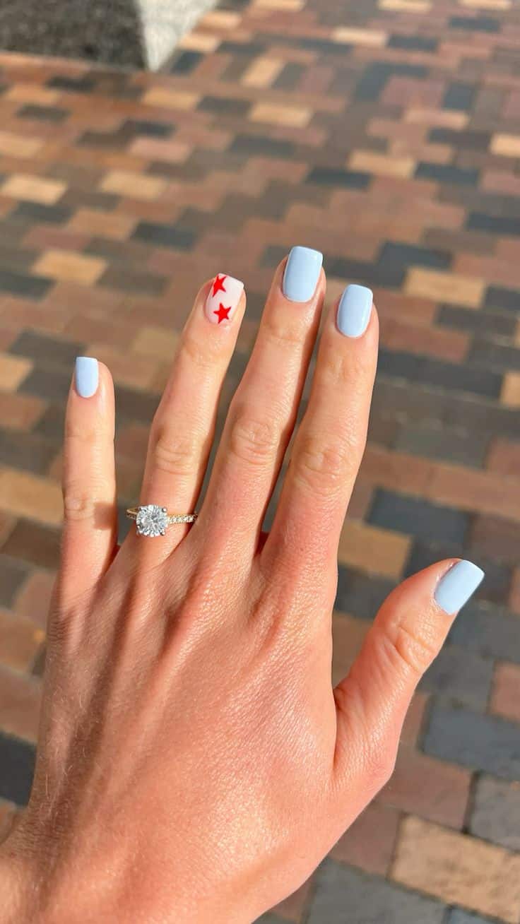 Short square nails painted in soft periwinkle blue with one sheer accent nail featuring two red stars, photographed outdoors on a brick background.