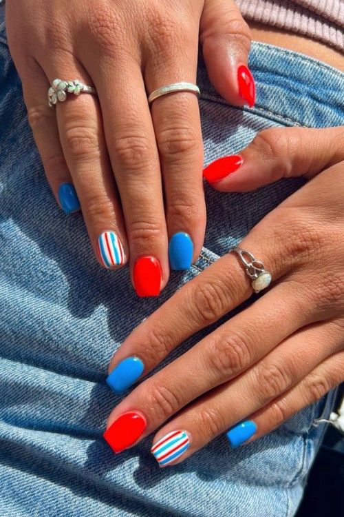 Short oval nails alternating between solid red and bright blue, with one white accent nail featuring thin red, white, and blue stripes, shown on two hands resting on denim.