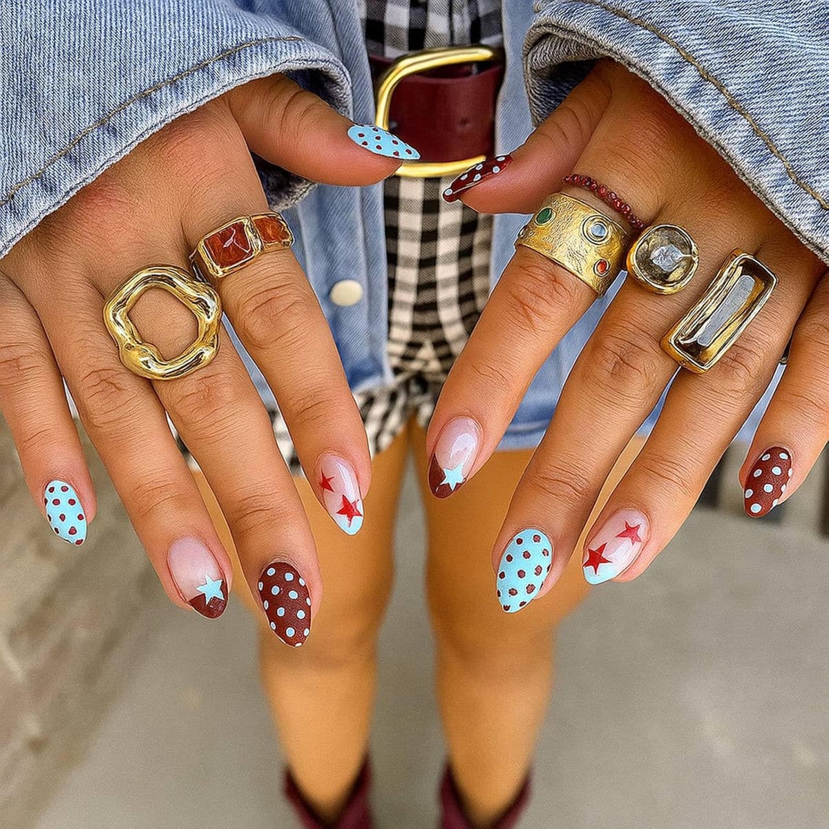 Medium almond nails featuring a retro-inspired patriotic design with sky blue polka dots on a brown base, red stars on a nude French tip, and mixed star and dot accents, styled with gold statement rings.