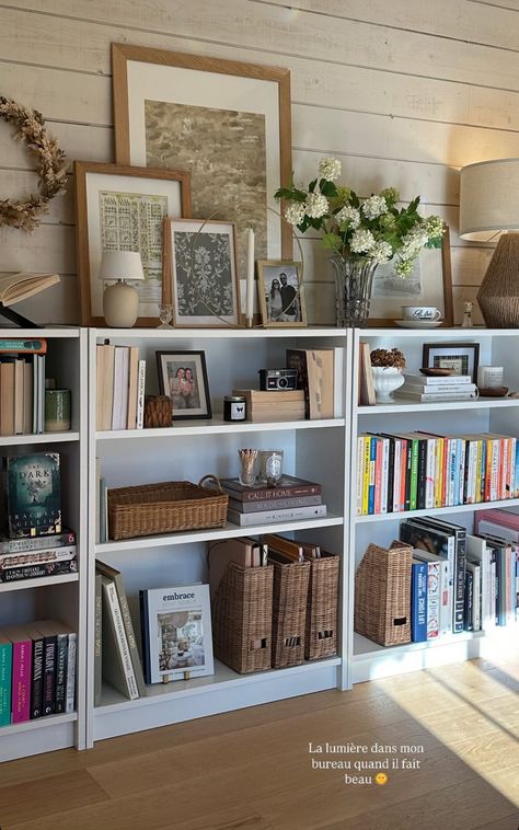 White bookshelves styled with wicker baskets, framed art, fresh flowers, and layered books in a warm home office