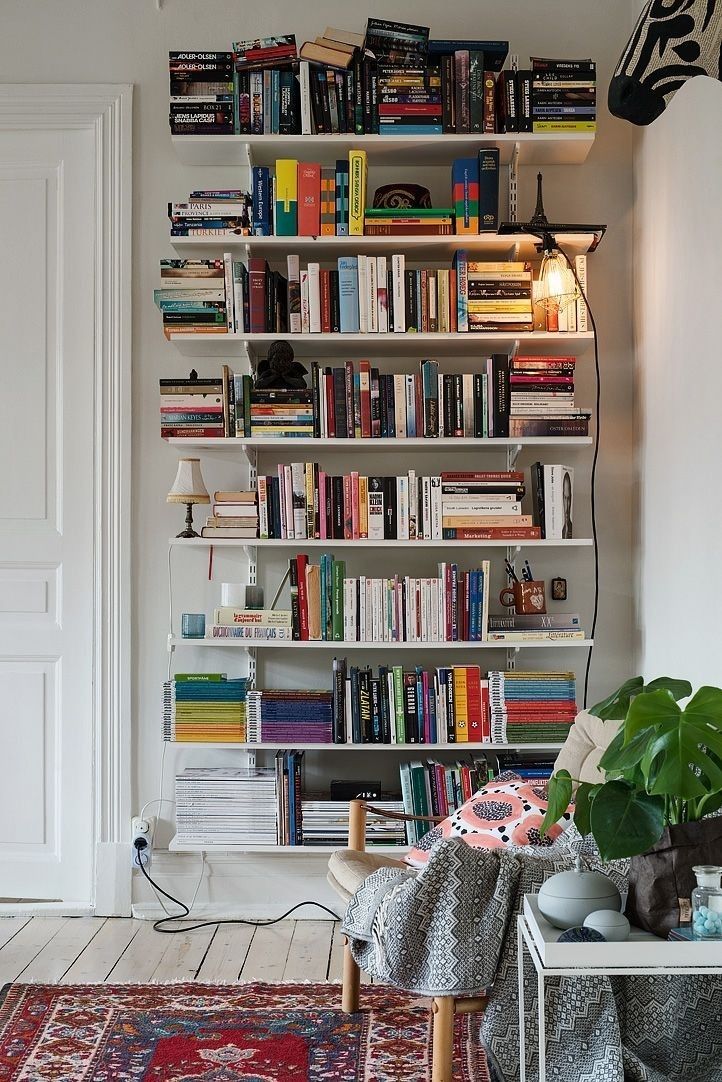 White wall-mounted bracket shelves packed with colourful books from top to bottom, with a monstera plant nearby