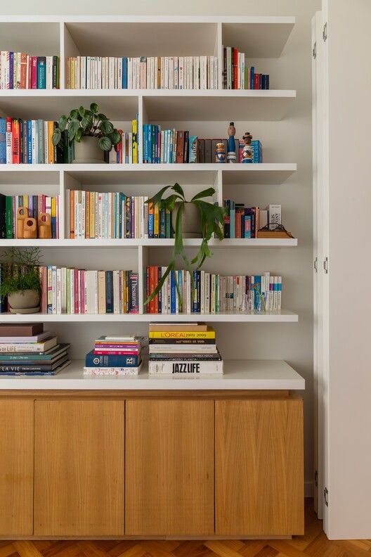 White built-in shelving unit with open shelves above and oak cabinet doors below, styled with colourful books and plants