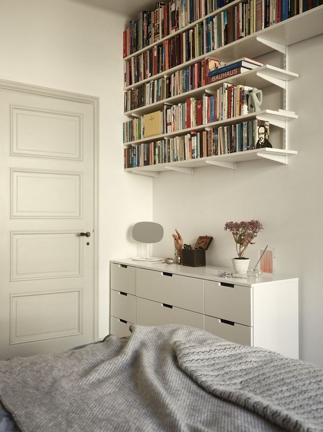 White wall-mounted shelves packed with books installed high above a light grey dresser in a minimalist white bedroom