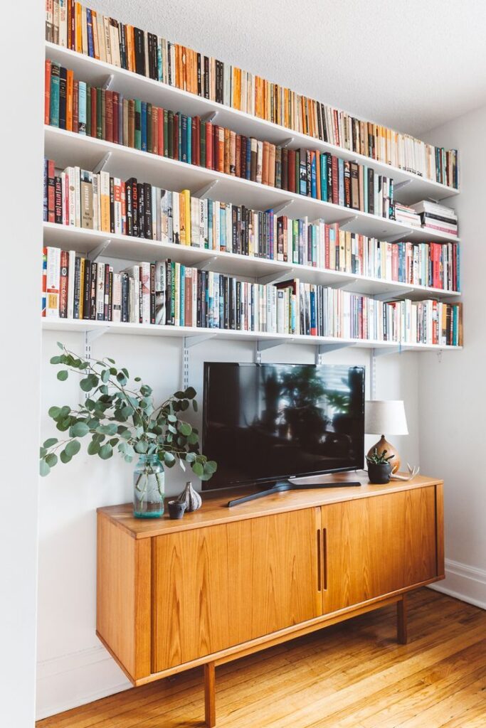 White wall-to-wall shelves above a mid-century oak sideboard, completely filled with books in a variety of colours