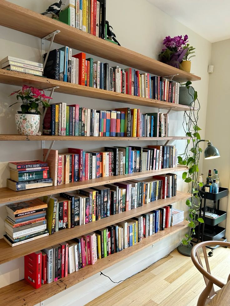 Wide natural timber floating shelves packed with books in a mix of colours, with a few small potted plants tucked in between