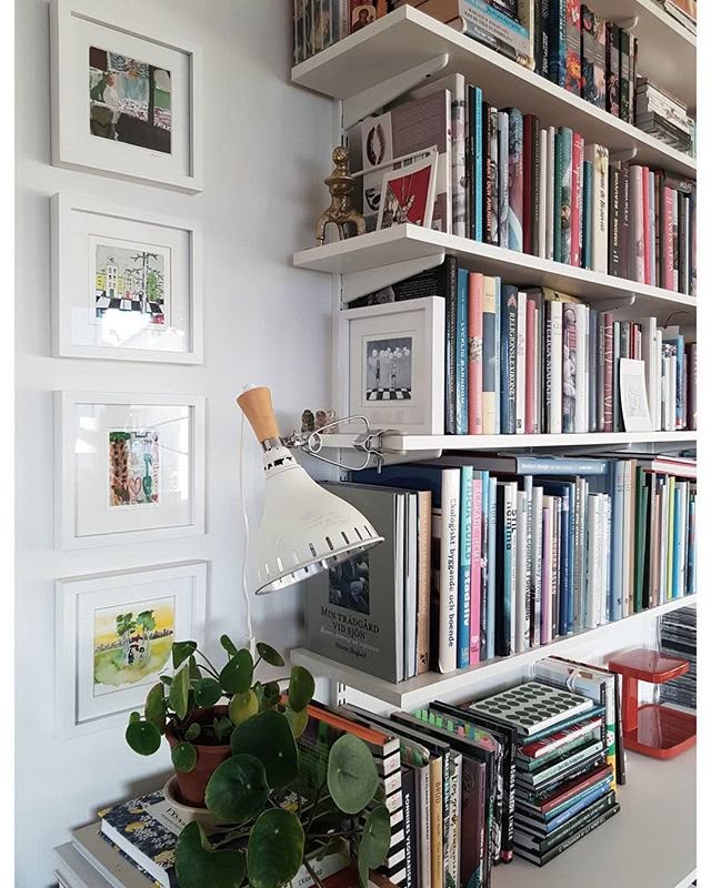 White floating shelves packed with books next to a small gallery wall of framed artworks, with a wall lamp and a pilea plant