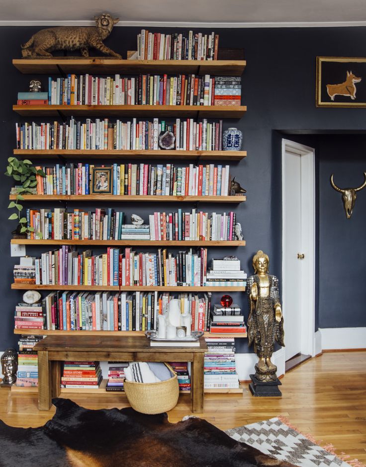 Floor-to-ceiling timber floating shelves packed with books and eclectic objects against a dark navy wall, with a brass statue and animal figurines