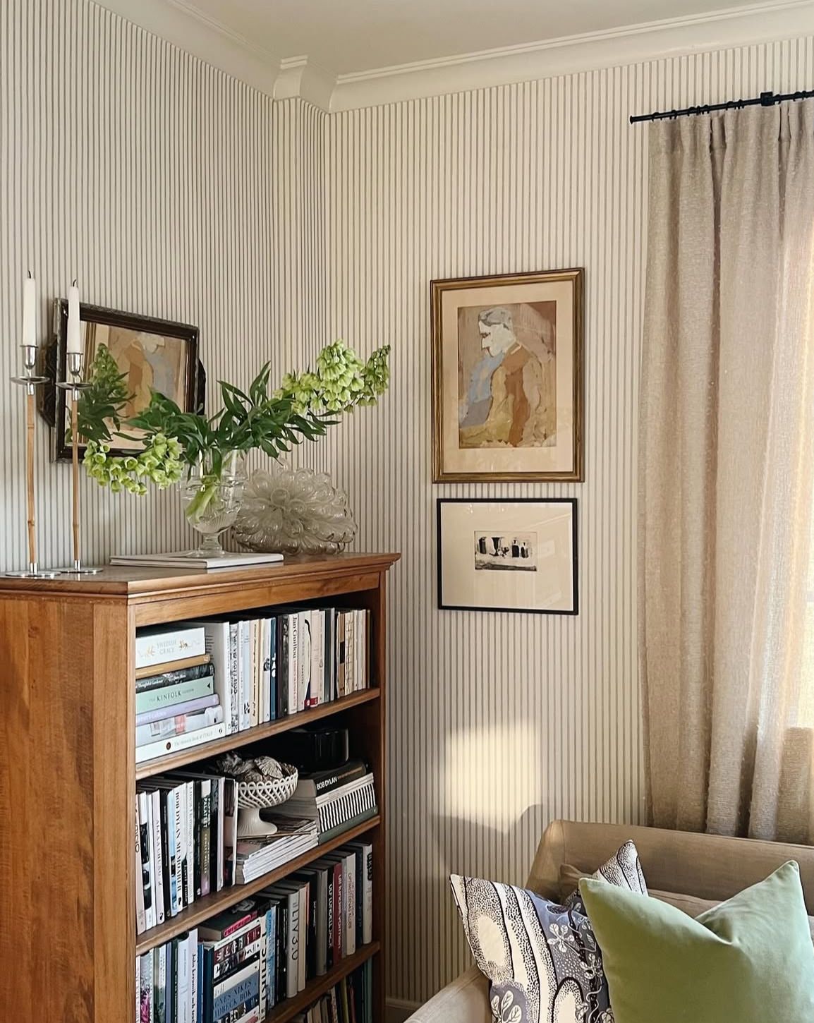 Top of a wooden bookcase styled with a glass vase of green flowers, a candle holder, and framed artwork against striped wallpaper