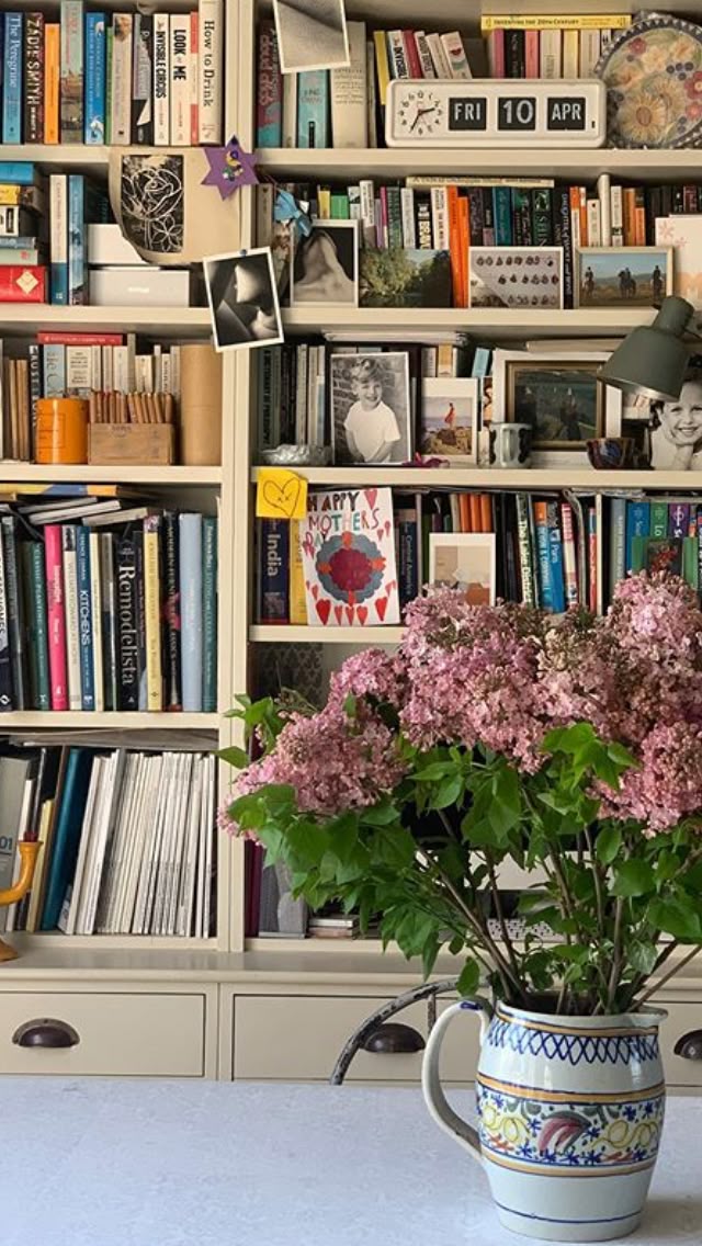 Cream built-in bookshelves filled with books, personal photos, postcards, and a ceramic jug of pink lilacs in the foreground