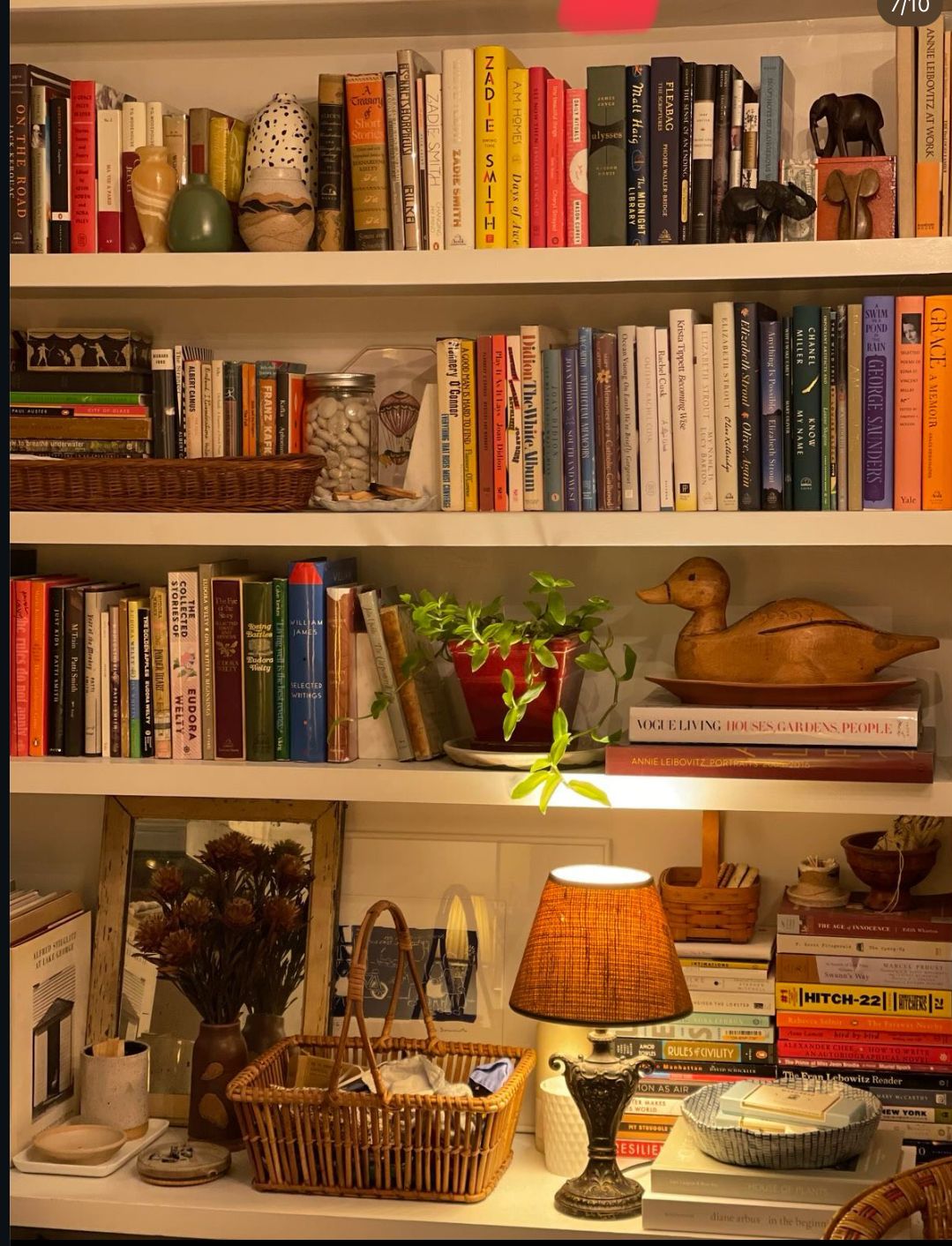 White bookshelves styled with a mix of colourful books, wicker baskets, a small plant, sculptural objects, and a glowing table lamp