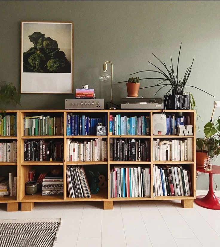 Natural wood low bookcase filled with books against a sage green wall, with a record player and plants on top