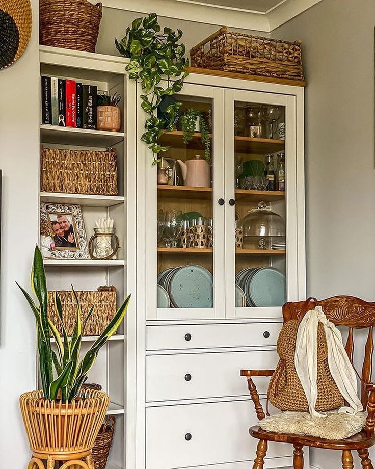White glass-fronted china cabinet styled with dusty blue plates, trailing plants, wicker baskets, and glassware next to an open wooden bookshelf.