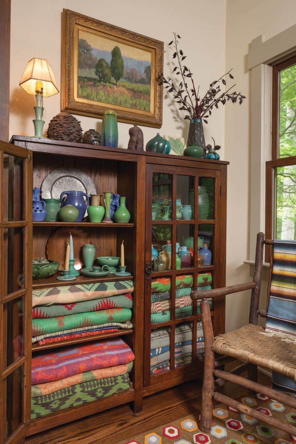 Dark wood glass-fronted cabinet filled with stacked colourful folded blankets on the lower shelves and green and blue handmade pottery on the upper shelves, topped with more ceramics and a vintage lamp in front of an oil painting.