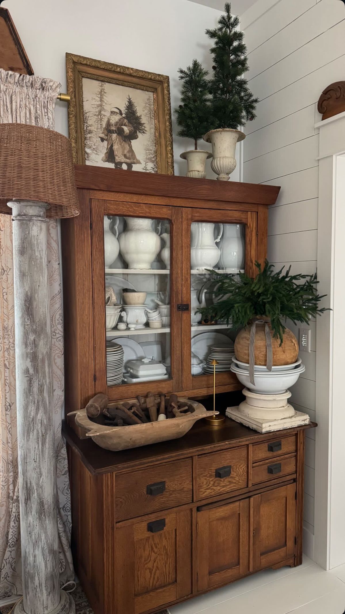 Antique golden oak hutch cabinet filled with white ironstone pitchers, stacked plates, and serving pieces, topped with a distressed white urn planter holding a small pine tree and a vintage Santa print in a gold frame.
