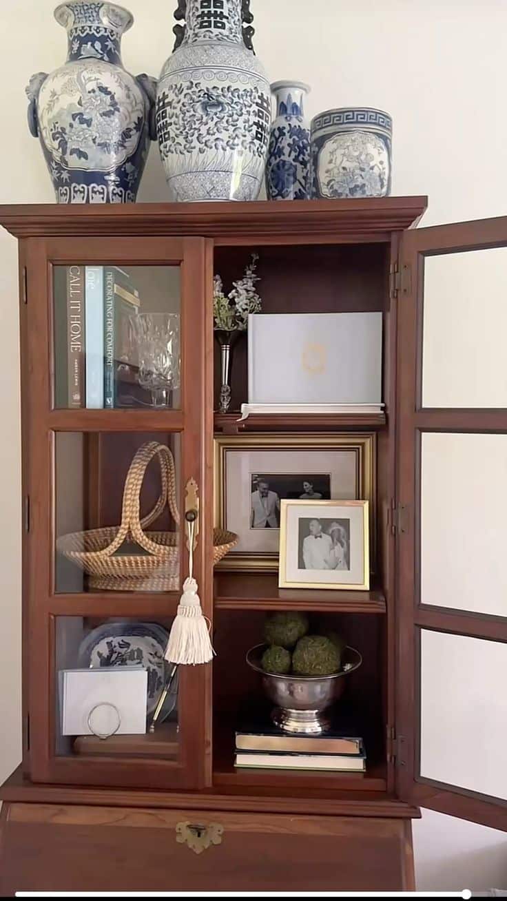 Warm timber secretary cabinet with glass doors open showing styled shelves with framed black and white wedding photos, a wicker basket, decorative books, and a moss bowl, topped with tall blue and white Chinese porcelain vases.