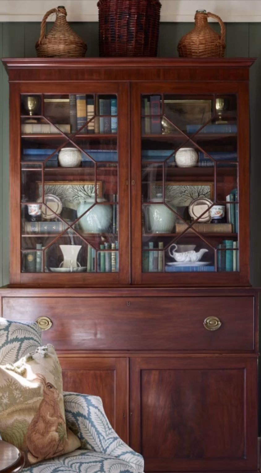 Antique mahogany china hutch with diamond-pane glass doors displaying antique books, pale blue and white ceramics, and white porcelain on wallpapered shelves, topped with wicker demijohn baskets.