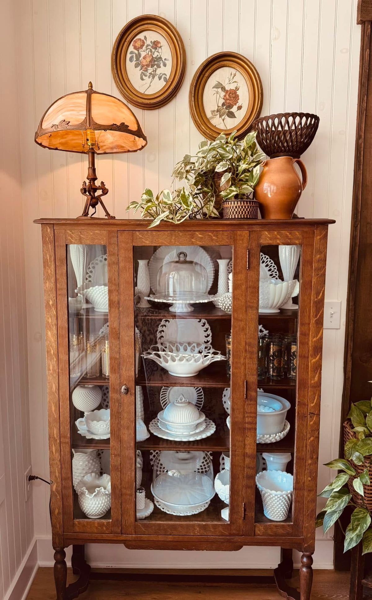 Antique golden oak china cabinet with turned legs fully stocked with a milk glass collection including hobnail vases, cake stands, bowls, and decorative dishes, topped with a vintage stained glass lamp and trailing plant.