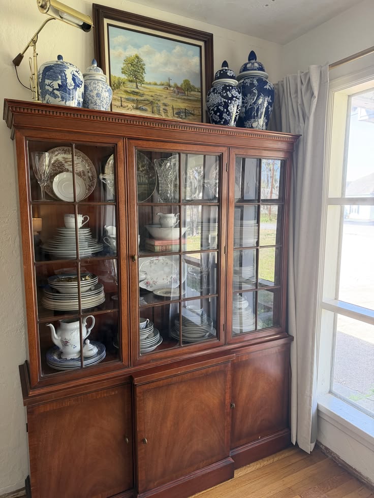 Large dark timber three-door china cabinet filled with mixed fine china, crystal glasses, and dinner sets, topped with oversized blue and white ginger jars and a framed landscape oil painting.