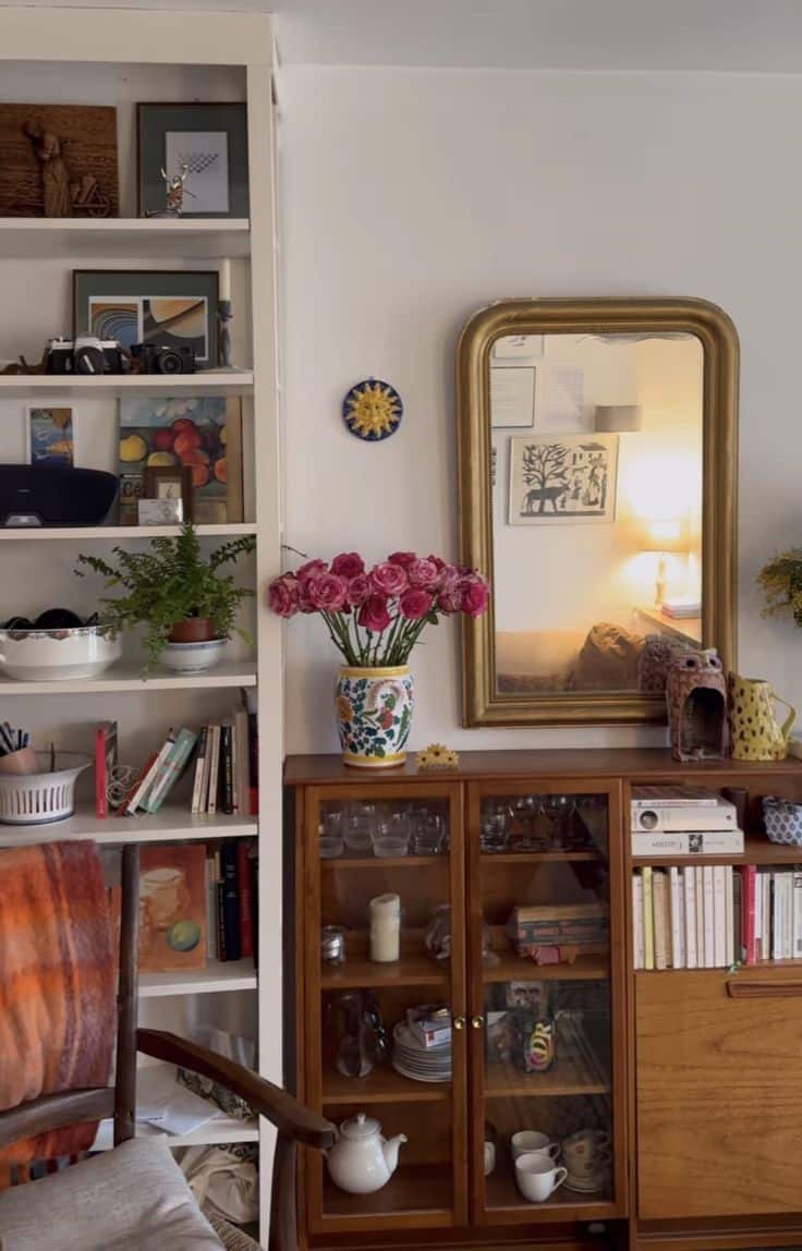 Small warm wood vintage glass-fronted cabinet in a living room displaying stacked plates, glassware, candles, and a white teapot, topped with a floral vase and gold-framed mirror on the wall above.