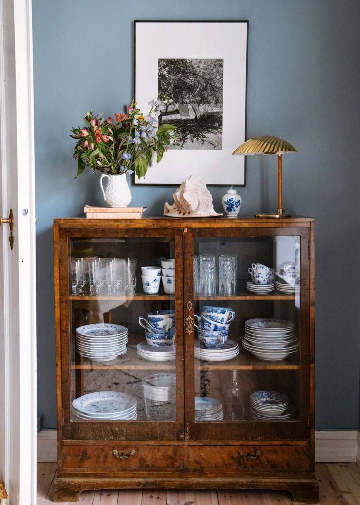 Low antique burled walnut glass-fronted cabinet displaying stacked blue and white china plates, cups, and saucers, topped with a white ceramic jug of flowers, stacked books, a brass mushroom lamp, and a small white ceramic.