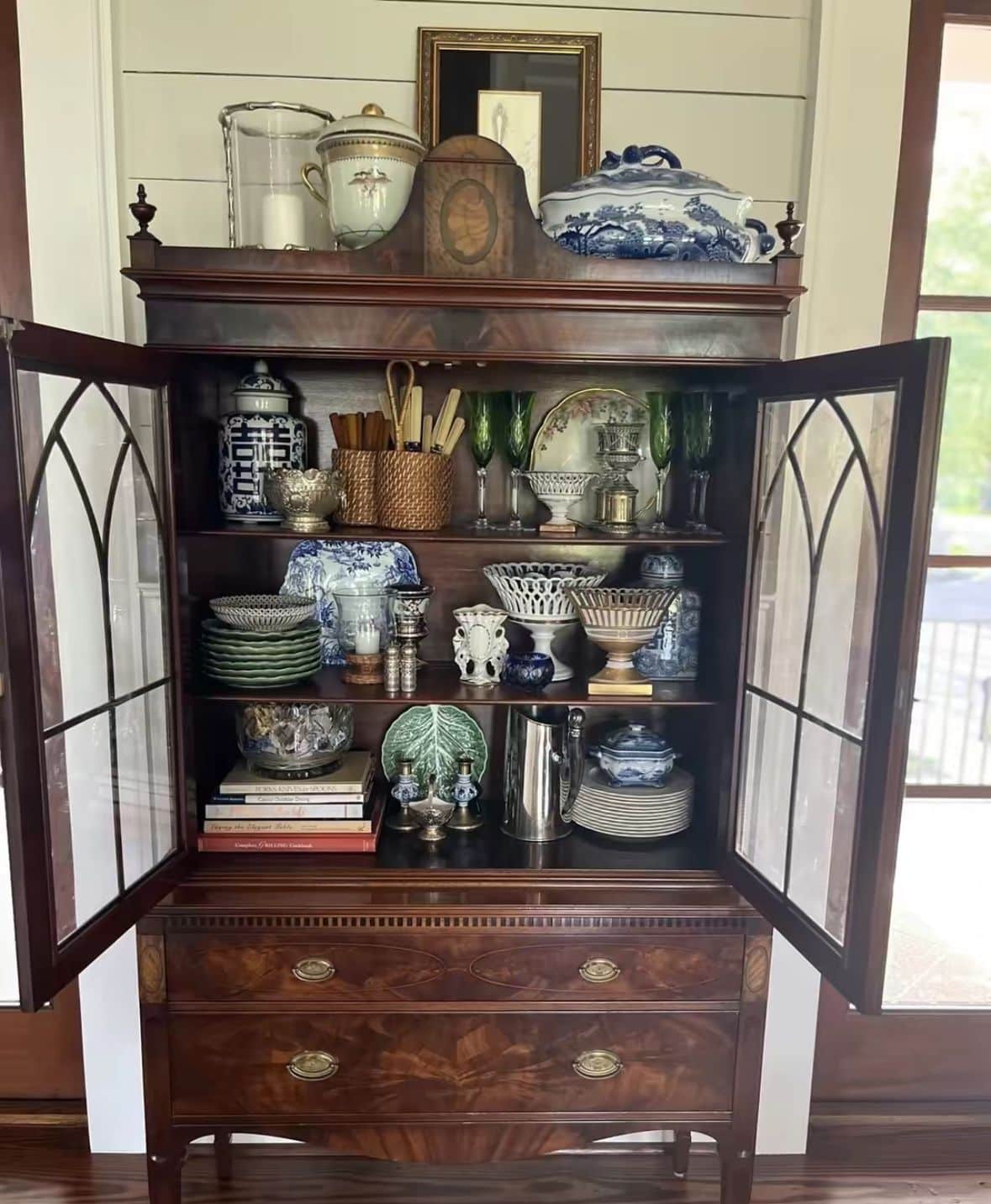 Open antique mahogany china cabinet with gothic arch glass doors displaying mixed blue and white ginger jars, green stacked plates, silver serving pieces, crystal glasses, and cookbooks, topped with large serving pieces and a framed print.