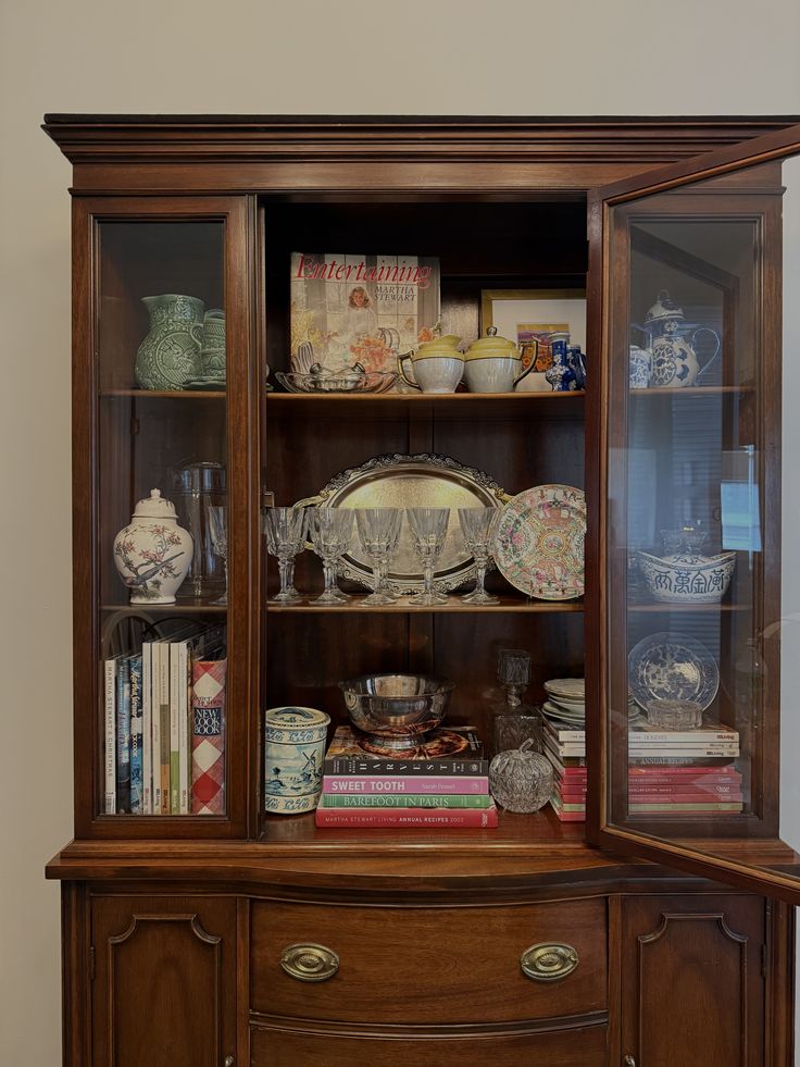 Dark timber china cabinet with curved glass sides open at the centre, displaying stacked cookbooks including a Martha Stewart Entertaining book, silver trays, crystal glasses, rose medallion plates, and mixed ceramics across three shelves.