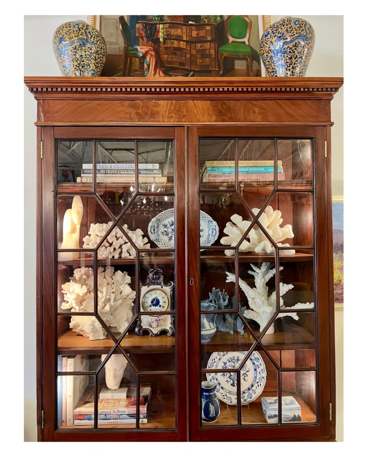 Antique mahogany china cabinet with diamond-pane glass doors displaying a curated collection of white coral specimens, blue and white china plates and ceramics, and antique books, topped with large blue and white ginger jars and a framed painting.