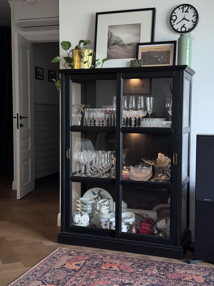 Black china cabinet with glass sliding doors displaying crystal glassware and fine china, topped with framed art, a plant, and a vintage clock.