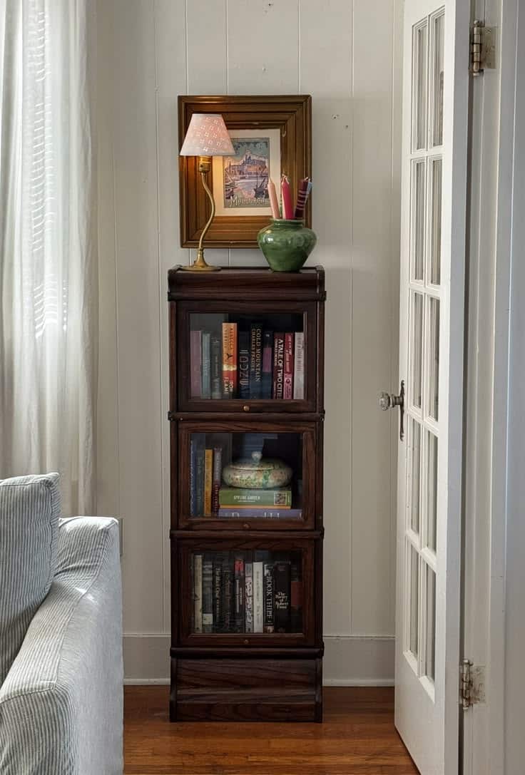 Dark wood stacked barrister bookcase filled with books and a decorative green ceramic vase on top, styled in a white-walled reading nook beside a French door.