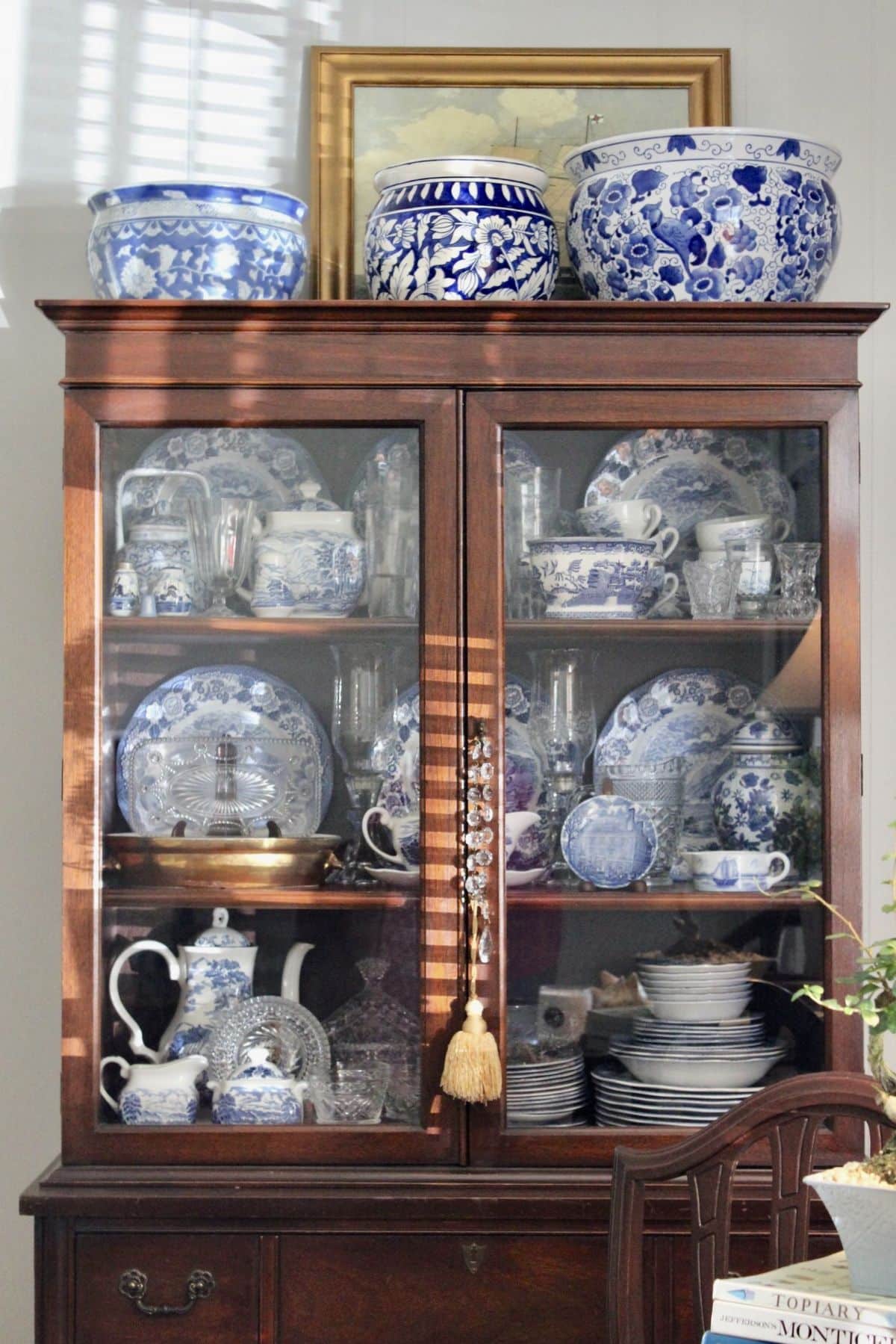 Dark timber china cabinet with glass doors fully styled with blue and white china pieces, plates, teapots, and jugs, topped with large blue and white ceramic fishbowl planters.