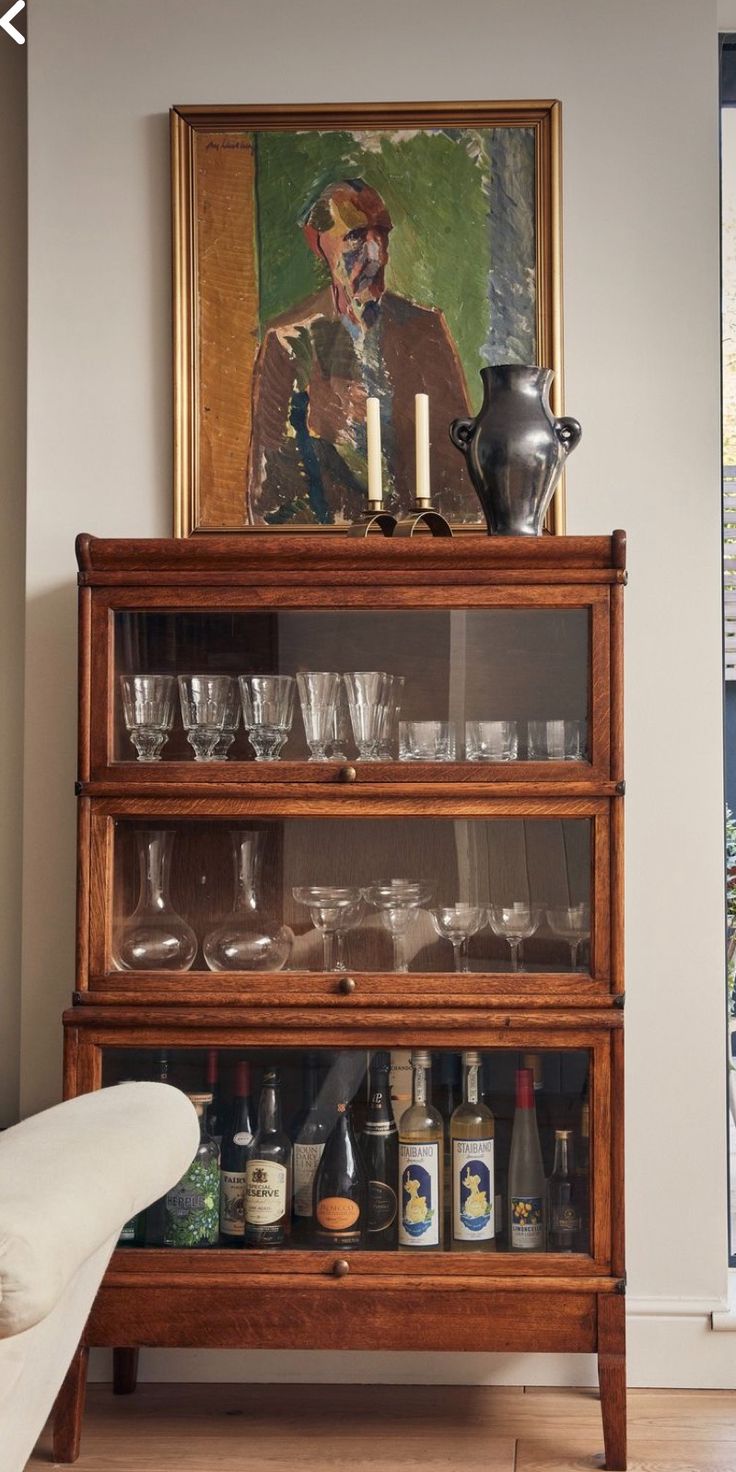 Warm oak barrister bookcase styled as a bar cabinet with glassware on upper shelves and wine bottles on the lower shelf, topped with a brass candleholder and dark ceramic jug in front of a painted portrait.