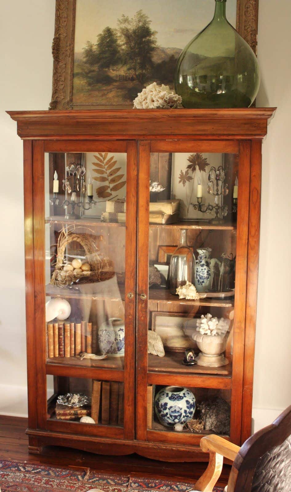 Antique warm timber glass-fronted cabinet displaying a curated collection of curiosities including coral, nests, antique books, candelabras, and blue and white ceramics, topped with a large green glass demijohn.