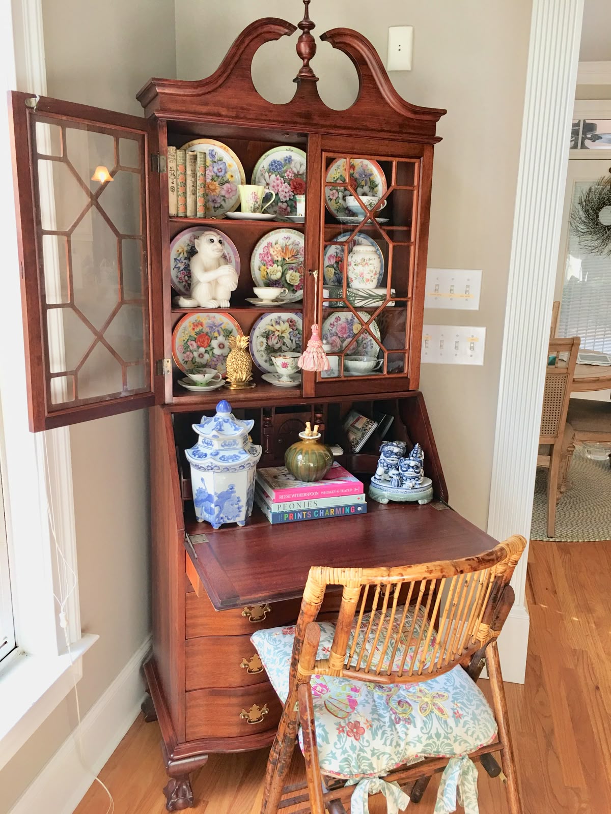 Antique mahogany secretary desk with glazed lattice-paned cabinet above displaying floral collector plates and a ceramic monkey figurine, with a rattan chair pulled up to the open desk surface.