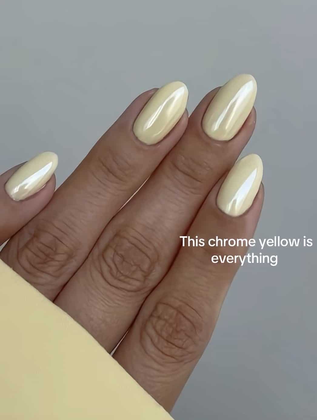 Almond nails in a glowing butter yellow chrome finish held against a grey background.