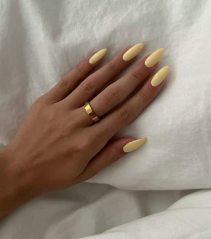 Long almond butter yellow nails on a hand resting on white bedding with a gold ring.