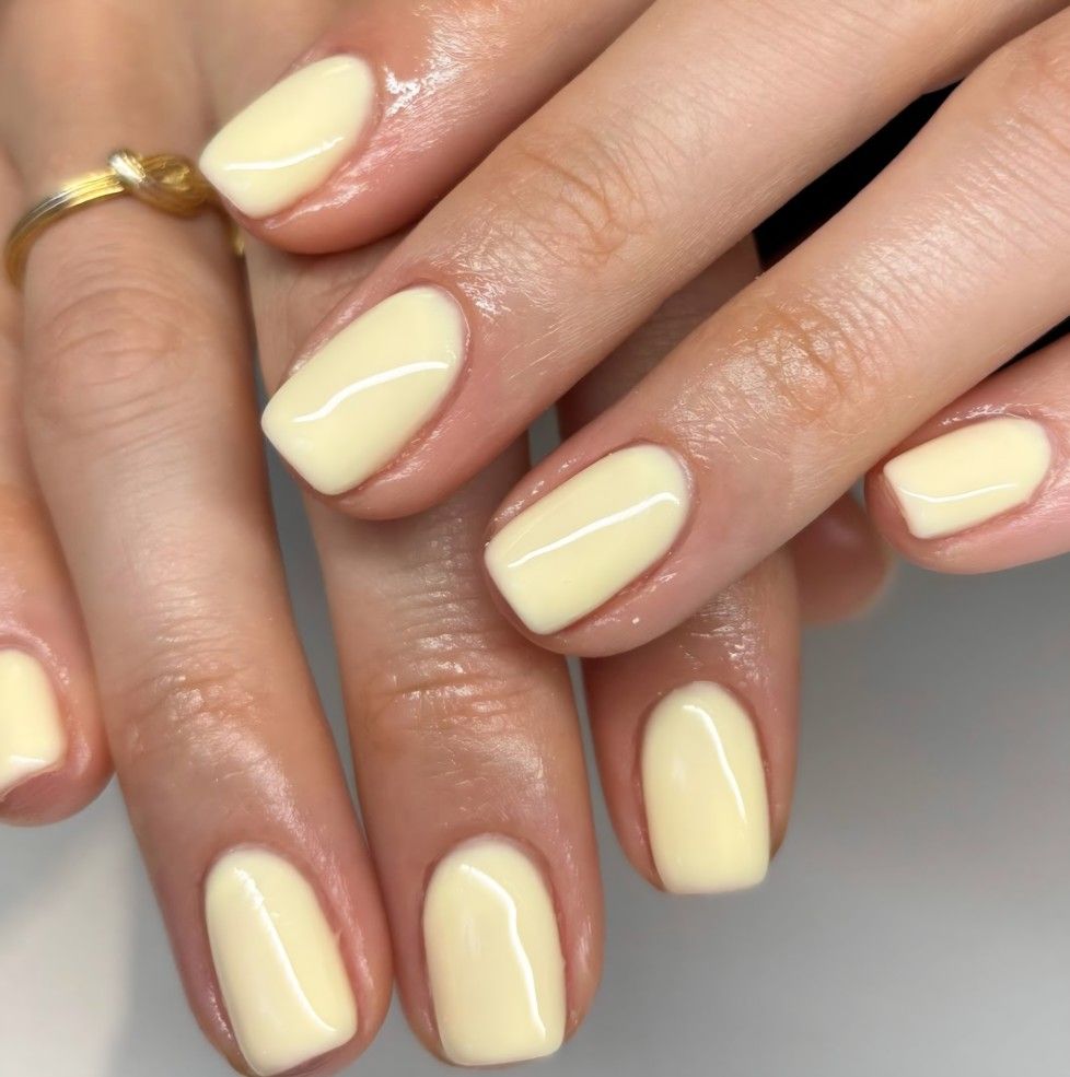 Short squoval nails in a glossy pale butter yellow with a thin gold ring.