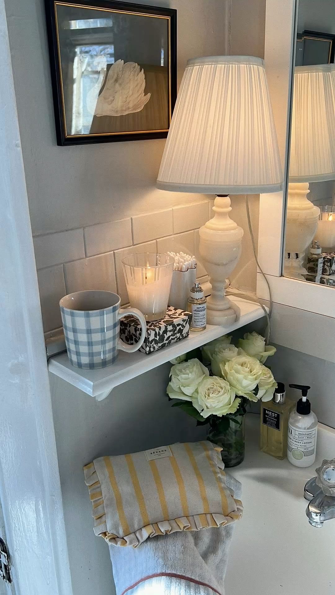 White bathroom shelf with a small lamp, candle, gingham mug and fresh white roses