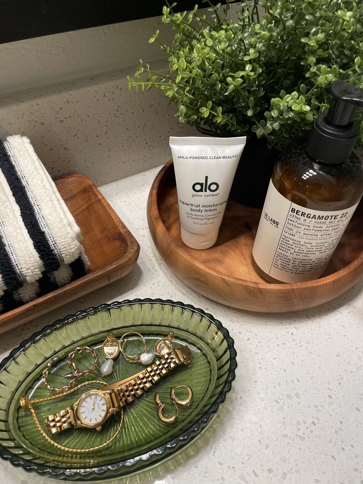Wooden tray with body lotion, a potted green plant and a green glass jewellery dish on a bathroom counter