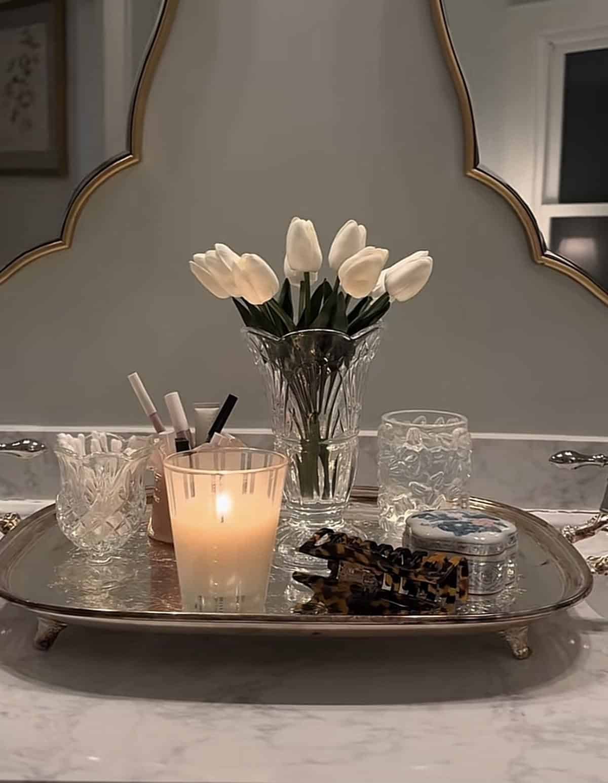 Footed silver tray styled with crystal glasses, white tulips, a candle and hair accessories on a marble counter